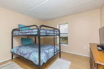 This bright bunk room offers a cozy setup with sturdy metal frames, matching blue bedding, soft rugs, and a dresser with TV—creating a practical, comfortable space ideal for kids, friends, or extra guests during your coastal getaway.