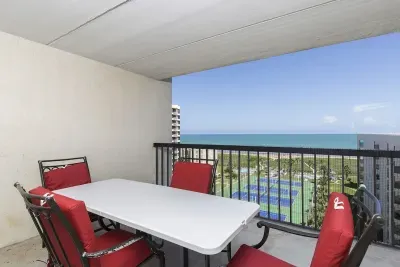 Relax on this spacious balcony featuring a crisp white dining set, vibrant red‑cushioned chairs, and sweeping coastal views overlooking tennis courts and the ocean—an inviting spot for morning coffee, sunset meals, and breezy downtime.