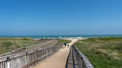 A peaceful boardwalk scene leading toward open sand and calm shoreline, offering an easy, unhurried start to any beach day. Soft light, natural textures, and a relaxed coastal flow set the tone for simple, effortless vacation moments.