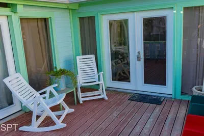 Cozy coastal porch featuring warm wood decking, classic white rocking chairs, and inviting double glass doors framed in soft mint. A small potted plant and relaxed outdoor setup create a charming spot to unwind and enjoy the breeze.
