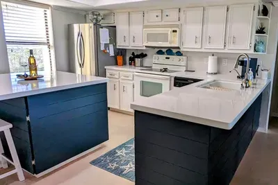 Modern kitchen with white upper cabinets, deep‑blue lowers, stainless appliances & bright countertops. The clean layout, natural light & coastal‑inspired accents create a fresh, guest‑ready space perfect for cooking, gathering & relaxed vacation.