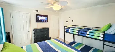 Coastal bedroom with a bed and matching bunk beds, all styled with striped blue‑and‑teal bedding. Wicker accents, seaside artwork and bright windows with turquoise curtains create a fresh, guest‑ready space perfect for families & relaxed stays.