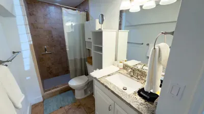 A spotless bathroom with a glass shower‑tub combo, granite countertop, and warm lighting. Thoughtfully organized with fresh towels and essentials, this space provides guests with comfort, cleanliness, and everyday convenience.