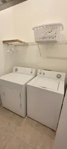 Bright, tidy laundry room featuring a full‑size washer and dryer, overhead shelving with storage, and clean tile flooring. A practical, easy‑to‑use space designed to keep guest stays convenient, organized, and stress‑free throughout their visit.