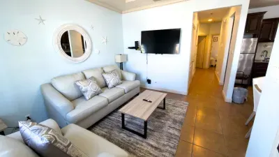 Bright, modern living space featuring cozy sofas, a sleek coffee table, and a stylish accent mirror. Enjoy an open layout flowing into the kitchen, soft lighting, and a welcoming setup ideal for relaxing, streaming, or gathering after a day out.