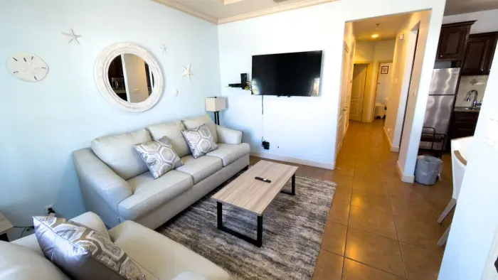 Bright, modern living space featuring cozy sofas, a sleek coffee table, and a stylish accent mirror. Enjoy an open layout flowing into the kitchen, soft lighting, and a welcoming setup ideal for relaxing, streaming, or gathering after a day out.