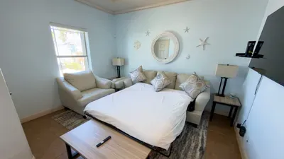 Comfortable sofa bed setup featuring a clean white sheet, geometric pillows, and warm bedside lighting. This flexible space offers guests a cozy, convenient sleeping option within a bright, thoughtfully arranged living area perfect for relaxing.