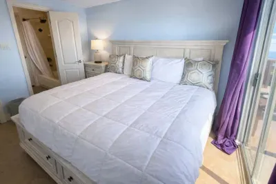 Bright, beachside bedroom retreat with comfy bed, geometric accent pillows, private bath access, and balcony views—sunlit space with purple drapes and coastal charm.