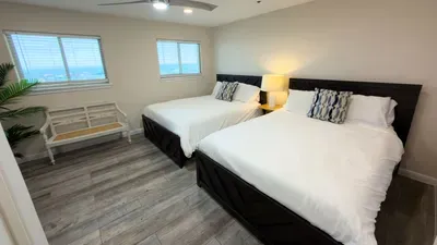 Twin Oceanview Beds Retreat
Share laughs or dreams in this breezy double-bedroom overlooking the sea. Crisp white bedding on dark frames, patterned pillows, lamp-lit nightstand, and potted palm add coastal flair.