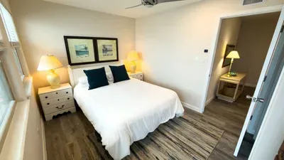Chic Coastal Bedroom Suite
Rest easy in this inviting bedroom with crisp white linens, vibrant teal pillows & golden lamps. Dual beach art prints adorn walls beside ample windows. Ceiling fan ensures comfort; adjacent door leads to bath.