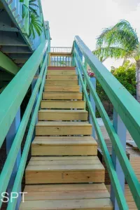 Sunny coastal stairway with light wood steps, green railings, and lush tropical plants. A bright, welcoming entry that sets the tone for a relaxed beach‑area stay.