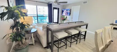 Spacious open‑concept living and dining area with a cozy sofa, high‑top table, modern décor, and large windows leading to a balcony with outdoor seating. A bright, inviting hub perfect for relaxing, gathering and enjoying effortless vacation comfort.