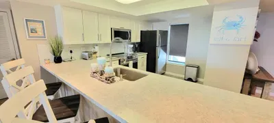 Bright, coastal‑styled kitchen featuring sleek white cabinetry, stainless appliances, a long breakfast‑bar counter with seating & warm decorative touches. A clean, functional space perfect for easy meal prep, casual dining & relaxed vacation living.