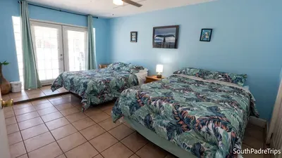 Bright, breezy retreat with tropical flair — unwind in this spacious coastal bedroom where sunlight filters through glass doors and soft blue tones meet leafy prints, creating a calm, island‑inspired escape steps from South Padre’s shoreline.