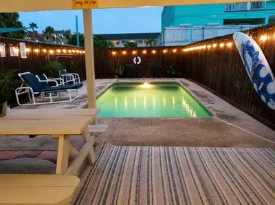 Cozy coastal hideaway with glowing pool and surf‑inspired charm. String lights shimmer over warm wood fencing as evening settles, inviting guests to unwind beneath the palms and soak in South Padre’s laid‑back island rhythm.