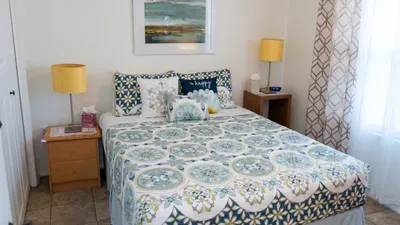 Serene Coastal Bedroom Retreat. Crisp white & blue floral quilt drapes plush king bed with patterned shams, "Happy" pillow & side tables holding lamps amid sea art, sheer drapes & tiled floors. Tranquil haven for dreamy beach nights!