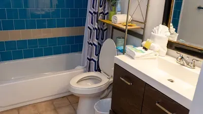 Crisp Ocean Blue Bathroom Retreat. Azure tiled tub/shower & modern toilet shine beside dark wood vanity stocked with plush towels, amenities & plant accents. Fresh, spa-ready space for refreshing post-beach rinses!