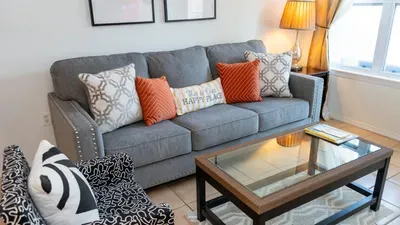 Sunny Coastal Living Nook w/ "Happy Place" Vibes. Gray sofa bursting with orange & white pillows including "Happy Place" welcomes lounging beside glass table, pattern chair & lamp-lit windows. Cheerful hub for relaxation & beachy gatherings!