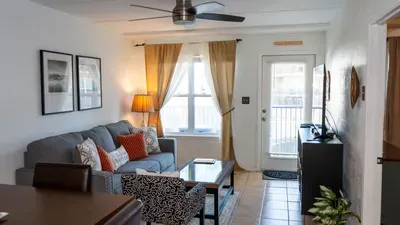 Cozy Gulf-View Living Area w/ Plush Seating. Charcoal sofa piled with pillows anchors sunlit nook beside glass coffee table, lamp-lit art & French doors to balcony. Pattern chair & console complete inviting coastal chill spot!