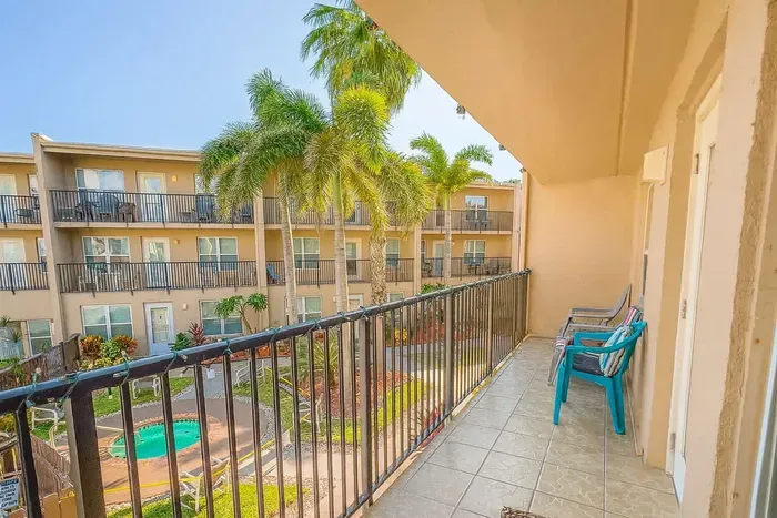 Private Resort Balcony Overlooking Palm-Lined Hot Tub Courtyard. Tan lounge chairs with colorful cushions perch on spacious balcony gazing down at bubbling spa, lush gardens, circle drive & tan condos amid swaying palms.