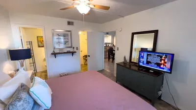 Inviting Coastal Bedroom Suite w/ Smart TV & Bath Access. Plush pink quilted bed anchors airy space with ceiling fan, dresser-mounted TV, nautical art, shelves & adjacent bath/kitchen doors. Cozy retreat for unwinding post-beach!