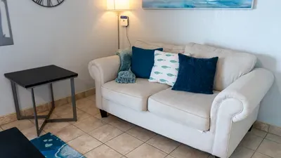 Relax in a bright, modern living room featuring crisp white sofas, coastal blue accents, and a sliding door opening to an ocean‑view balcony. A stylish, airy space perfect for unwinding, gathering, and enjoying effortless vacation comfort.