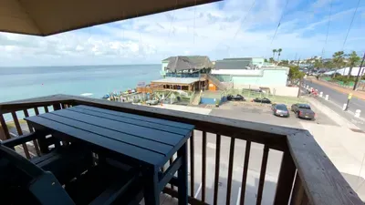 Relax on a breezy coastal balcony overlooking calm blue waters, lively beachfront dining, and a vibrant shoreline scene. Perfect for morning coffee or sunset unwinding, this inviting spot blends comfort, scenery, and effortless vacation vibes.
