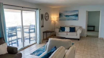 Coastal Living Room Glow — A bright living room with crisp white sofas, blue accents, and a sliding door opening to an ocean‑view balcony creates the perfect space for relaxing, gathering, and enjoying breezy coastal comfort.
