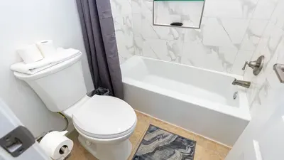 Bright, well-kept bathroom featuring a clean tub with marble‑style tile surround, organized shelving, & soft neutral tones. Practical touches like fresh towels, paper & a cozy bath mat create a simple, comfortable space for good guest convenience.