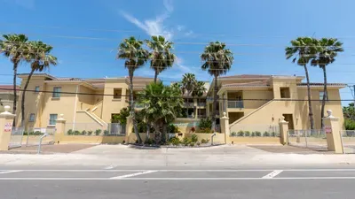 Inviting coastal-style building featuring light stucco exteriors, dual staircases, and private balconies framed by tall palms. Bright, sunny surroundings and a well-kept façade create a welcoming first impression for a relaxed, beach‑area stay.