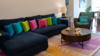Enjoy comfort and vibrant coastal charm in the living room with a deep blue sectional, cheerful pillows, whimsical sea turtle accents, and a cozy reading chair—perfect for relaxing and unwinding after fun-filled beach days.