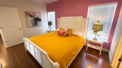 Cheerful bedroom retreat with a plush yellow comforter, flamingo pillow, pink accent wall, and whimsical lamps. Large windows fill the room with light—perfect for relaxing and waking up refreshed on vacation.