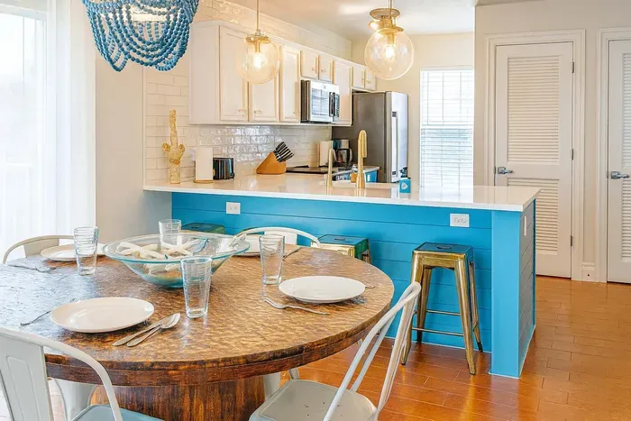 Bright and stylish dining space with a rustic round table for four, chic turquoise accents, and a modern kitchen with stainless appliances and breakfast bar seating—ideal for relaxing meals and gathering with friends or family.