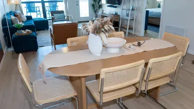 Bright dining area with woven‑back chairs, light wood table, and soft natural décor, opening into a cozy living space and bedroom, offering guests an inviting, well‑designed layout perfect for shared meals, relaxing moments, and a comfortable stay.