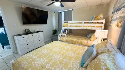 Relax in this spacious coastal bedroom featuring a full bunk, plush queen bed, soft lighting, and cheerful décor—an inviting setup ideal for families, shared spaces, and comfortable vacation downtime in a bright, welcoming atmosphere.