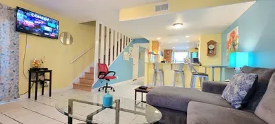 Modern, inviting living room with a cozy sectional, vibrant accents, and easy access to the patio for fresh coastal air. The open kitchen, breakfast bar seating, and warm natural light create a comfortable, social space ideal for a carefree stay.