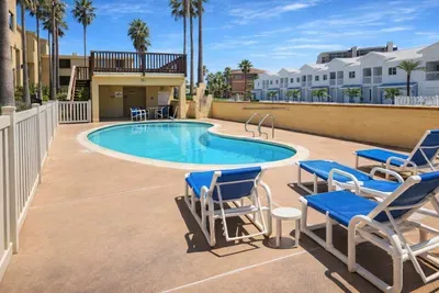 Modern poolside retreat with tropical vibes—kidney-shaped pool, sun loungers, palm trees, and shaded deck seating just steps from white townhomes and beach access. Ideal for relaxing getaways and family vacations.
