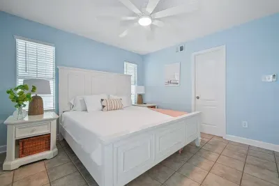 Serene main-level king bedroom with coastal décor, ceiling fan, smart TV, private balcony access, and a connected ensuite bathroom—perfect for guests seeking comfort, privacy, and easy access to the home’s main living areas.