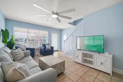 Spacious main-level living room with plush seating, smart TV, ceiling fan, and large windows for natural light. Conveniently located near the stairs and balcony access—ideal for relaxing, movie nights, or gathering after a day at the beach.
