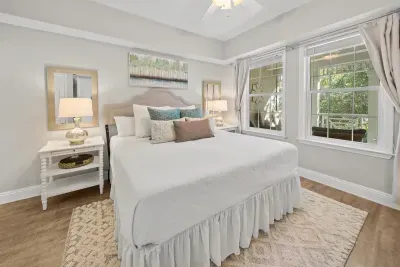 A restful bedroom with soft white bedding, a bedside lamp and big windows. A calm place to land after a full day on 30A.