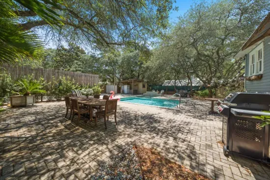 The backyard is built for gathering — a paver patio with outdoor dining, a Weber grill, and the heated pool just beyond, all framed by Grayton's signature live oaks.