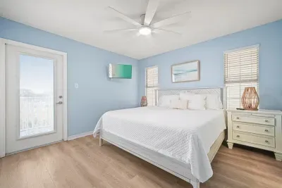 Spacious primary bedroom on the Unit B side featuring a king bed, private ensuite bathroom, and a walkout balcony with bay views. Bright, coastal design with TV, ceiling fan, and direct access to outdoor seating.