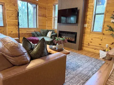 Cozy cabin living room with fireplace, TV, and bright woodland views