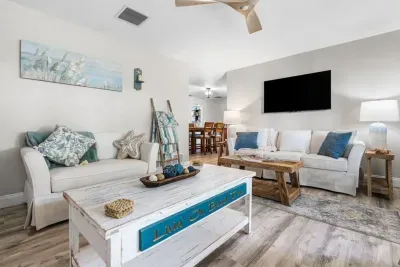 A bright coastal living room at Twin Oaks Coastal Luxury with white sofas, a distressed `Livin` On Beach Time` coffee table, beach grass artwork, and a wall-mounted Smart TV — the perfect group hangout space.