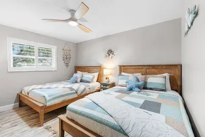 Castaway's Respite, a serene double-queen bedroom at Twin Oaks Coastal Luxury, features coastal patchwork quilts, seashell wall art, warm wood headboards, and a soft, beachy feel that sleeps four comfortably.