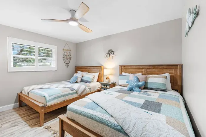 Castaway's Respite, a serene double-queen bedroom at Twin Oaks Coastal Luxury, features coastal patchwork quilts, seashell wall art, warm wood headboards, and a soft, beachy feel that sleeps four comfortably.