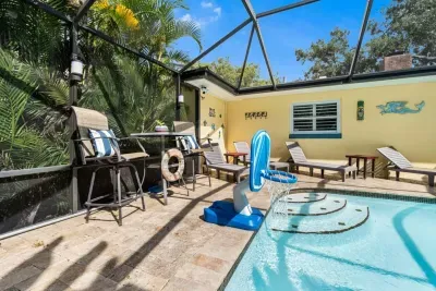 Sun loungers and a high-top bistro set line the sunny pool deck at Twin Oaks Coastal Luxury, complete with a pool basketball hoop — a full resort experience in your private screened backyard.