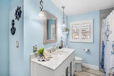 A charming nautical-themed shared bathroom with sky-blue walls, a marble-top vanity, ship wheel and anchor décor, and a tiled shower — fully stocked with complimentary toiletries at Twin Oaks Coastal Luxury.