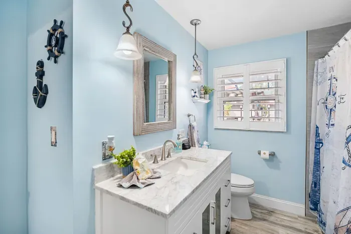 A charming nautical-themed shared bathroom with sky-blue walls, a marble-top vanity, ship wheel and anchor décor, and a tiled shower — fully stocked with complimentary toiletries at Twin Oaks Coastal Luxury.
