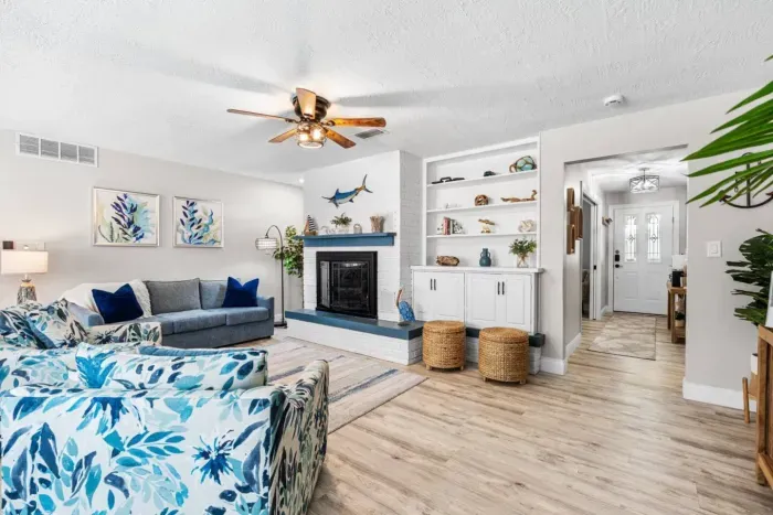 The main living room at Twin Oaks Coastal Luxury, showcasing the white brick fireplace with blue accents, built-in shelving, bold coastal floral armchairs, and a clear sightline to the bright entryway and front door.